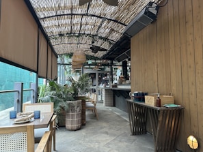 A cozy outdoor cafe with wooden furniture and a bamboo awning creates a serene ambiance. Potted plants add greenery, while tables are set with napkins and candles. A small, organized bar is visible in the background.
