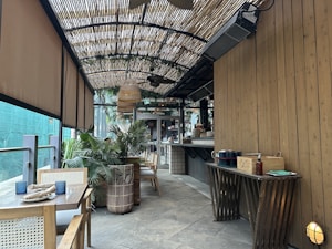 A cozy outdoor cafe with wooden furniture and a bamboo awning creates a serene ambiance. Potted plants add greenery, while tables are set with napkins and candles. A small, organized bar is visible in the background.