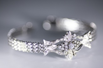 A close-up view of a delicately crafted bracelet featuring an intricate design with sparkling diamonds and detailed metalwork. The bracelet has a woven pattern and ornate central motif resembling leaves and flowers. The background is softly blurred creating a bokeh effect, highlighting the elegance of the jewelry.