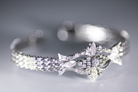 A close-up view of a delicately crafted bracelet featuring an intricate design with sparkling diamonds and detailed metalwork. The bracelet has a woven pattern and ornate central motif resembling leaves and flowers. The background is softly blurred creating a bokeh effect, highlighting the elegance of the jewelry.