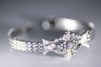 A close-up view of a delicately crafted bracelet featuring an intricate design with sparkling diamonds and detailed metalwork. The bracelet has a woven pattern and ornate central motif resembling leaves and flowers. The background is softly blurred creating a bokeh effect, highlighting the elegance of the jewelry.