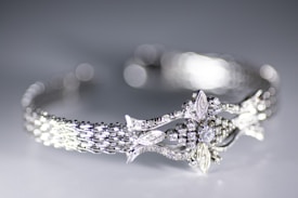 A close-up view of a delicately crafted bracelet featuring an intricate design with sparkling diamonds and detailed metalwork. The bracelet has a woven pattern and ornate central motif resembling leaves and flowers. The background is softly blurred creating a bokeh effect, highlighting the elegance of the jewelry.