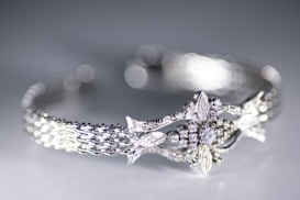 A close-up view of a delicately crafted bracelet featuring an intricate design with sparkling diamonds and detailed metalwork. The bracelet has a woven pattern and ornate central motif resembling leaves and flowers. The background is softly blurred creating a bokeh effect, highlighting the elegance of the jewelry.