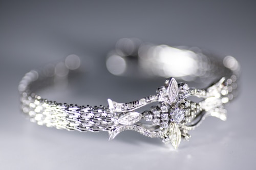 A close-up view of a delicately crafted bracelet featuring an intricate design with sparkling diamonds and detailed metalwork. The bracelet has a woven pattern and ornate central motif resembling leaves and flowers. The background is softly blurred creating a bokeh effect, highlighting the elegance of the jewelry.