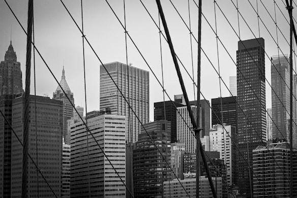 A cityscape featuring tall skyscrapers fills the background, creating a dense urban atmosphere. The foreground displays structural cables, likely from a bridge, intersecting the view of the buildings and adding an element of industrial design.