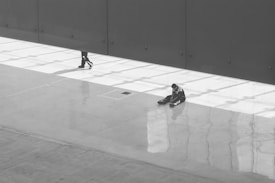 A vast, mostly empty space with smooth, reflective flooring and stark lighting. A solitary figure sits on the ground in the right part of the image, while only the legs of another person are visible as they walk by, obscured by a large vertical structure casting shadows.