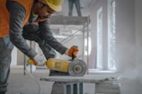 A construction worker is operating a circular saw to cut a concrete block inside a partially constructed building. The worker is wearing a hard hat, an orange safety vest, and protective gloves. Scaffolding and unfinished walls can be seen in the background, along with some dust and construction debris on the floor.