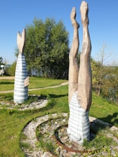 Two wooden sculptures resembling human forms are placed outdoors on a grassy area. One sculpture depicts a figure standing upright, while the other is inverted, showing legs and feet directed towards the sky. The artwork is surrounded by stone circles and small white pebbles, with trees and a clear blue sky in the background.