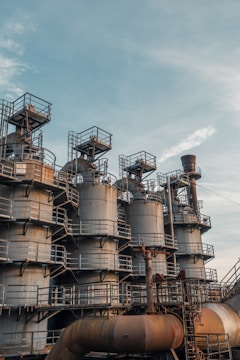 A series of large industrial tanks with metal railings and stairs are positioned against a backdrop of a clear sky. The structures are metallic, showing signs of rust and weathering. The intricate network of pipes and ladders suggests an industrial or manufacturing setting.