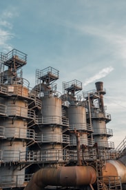 A series of large industrial tanks with metal railings and stairs are positioned against a backdrop of a clear sky. The structures are metallic, showing signs of rust and weathering. The intricate network of pipes and ladders suggests an industrial or manufacturing setting.