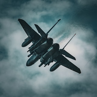 A military fighter jet soars through a cloudy sky, showcasing its sleek design and power. The aircraft is angled sharply, emphasizing its aerodynamic shape against the backdrop of ominous gray clouds.