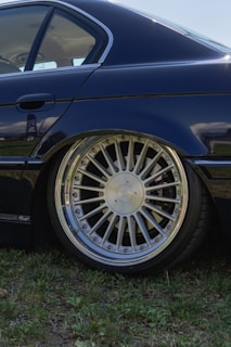 A close-up view of a car with a shiny, detailed alloy wheel. The vehicle appears to be parked on grassy terrain, and the sun reflects off the body and wheel. The car's sleek, dark paint contrasts with the surroundings.