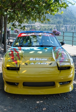 A customized yellow car with vibrant decals on the hood, featuring artistic flame designs. The windshield displays various stickers, and the car is parked by a scenic lake with mountains in the background. Surrounding trees and flowering plants add a natural ambiance.