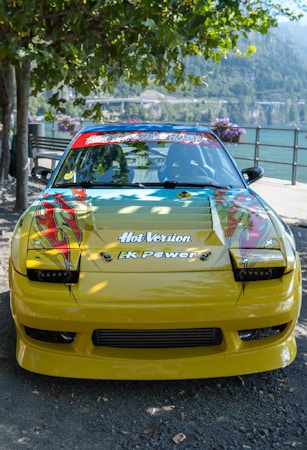 A customized yellow car with vibrant decals on the hood, featuring artistic flame designs. The windshield displays various stickers, and the car is parked by a scenic lake with mountains in the background. Surrounding trees and flowering plants add a natural ambiance.