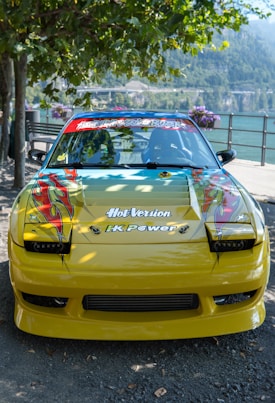A customized yellow car with vibrant decals on the hood, featuring artistic flame designs. The windshield displays various stickers, and the car is parked by a scenic lake with mountains in the background. Surrounding trees and flowering plants add a natural ambiance.