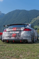 A modified sports car is parked on a grassy area with a backdrop of lush green mountains. The car features a custom paint job with a red stripe, a large rear spoiler, and quad exhausts.