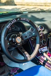 An interior view of a car featuring a custom steering wheel labeled 'Fatlace Vertex' with a graphic in the center. The dashboard includes several round gauges, and the upholstery exhibits colorful, patterned designs. The gear shift and surrounding console are vibrantly decorated.