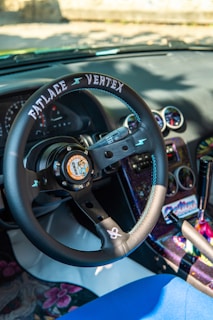 An interior view of a car featuring a custom steering wheel labeled 'Fatlace Vertex' with a graphic in the center. The dashboard includes several round gauges, and the upholstery exhibits colorful, patterned designs. The gear shift and surrounding console are vibrantly decorated.