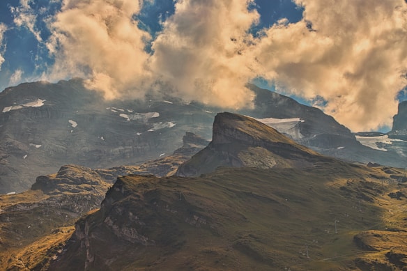 A dramatic mountain landscape under a sky filled with large, fluffy clouds. The mountain peaks have patches of snow, and the slopes are covered in green grass. The lighting creates a contrast between the shadows and sunlit areas, emphasizing the ruggedness of the terrain.