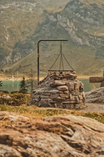 A stone fire pit with a metal frame above it, located in a mountainous landscape. The background features rocky hills and a body of water, surrounded by sparse vegetation and trees.