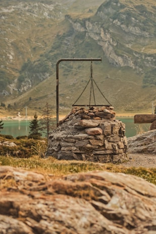 A stone fire pit with a metal frame above it, located in a mountainous landscape. The background features rocky hills and a body of water, surrounded by sparse vegetation and trees.