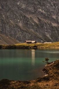 A peaceful lakeside scene with a clear, turquoise lake in the foreground. Two buildings, possibly cabins, sit on a small strip of grass surrounded by majestic, steep, rocky mountains in the background.