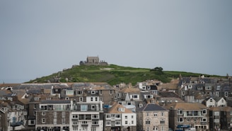 A coastal village with rows of traditional stone buildings topped with slate roofs extends below a green hill. Atop the hill, a small stone structure offers expansive views, surrounded by grass and a few shrubs.