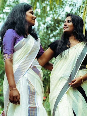 Two women dressed in traditional attire are standing outdoors, engaging in conversation while smiling at each other. The background features lush green vegetation, suggesting a natural, scenic setting.