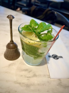 A glass filled with a refreshing beverage containing lime slices and fresh mint leaves, accompanied by a metallic decorative item and a napkin with a logo on a marble tabletop. A red straw is placed in the drink.