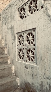 A rustic concrete wall features decorative geometric designs with circular patterns and star-shaped cutouts. Next to the wall, cracked concrete steps lead upward, suggesting an aged or abandoned environment.