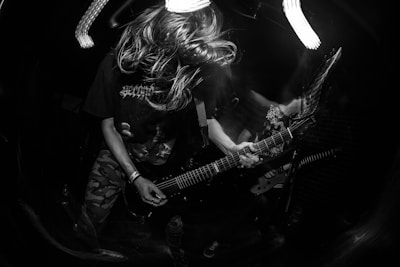 A black and white photo captures a dynamic scene of a long-haired guitarist intensely playing on stage. The motion blur accentuates the energy and movement of the musician, who is wearing a dark t-shirt and camouflage pants. The scene is dimly lit, highlighting the vibrant performance.