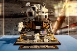 A detailed LEGO model of the Apollo Lunar Lander is displayed, featuring intricate design and attention to detail. The model includes small astronaut figures and the United States flag, all set on a blue surface against a moon-like background.