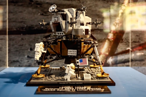 A detailed LEGO model of the Apollo Lunar Lander is displayed, featuring intricate design and attention to detail. The model includes small astronaut figures and the United States flag, all set on a blue surface against a moon-like background.