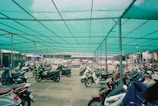 A sheltered parking area filled with numerous parked motorcycles, featuring a teal shade cloth overhead supported by metal poles. In the background, a row of parked cars is visible, with urban buildings partially seen beyond the parking area.