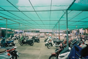 A sheltered parking area filled with numerous parked motorcycles, featuring a teal shade cloth overhead supported by metal poles. In the background, a row of parked cars is visible, with urban buildings partially seen beyond the parking area.