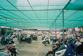 A sheltered parking area filled with numerous parked motorcycles, featuring a teal shade cloth overhead supported by metal poles. In the background, a row of parked cars is visible, with urban buildings partially seen beyond the parking area.