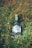 A bottle of gin is placed among tall green grass and small yellow wildflowers. The bottle has a wooden cork and a hexagonal label reading 'Anaë Gin de France BIO'. The glass is clear with a textured design, reflecting light.