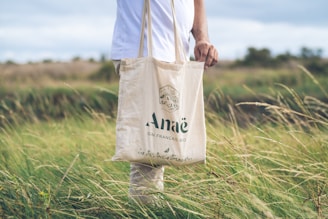 A person is standing in a field holding a beige tote bag with the logo and text 'Anaë Gin Francais Bio' printed on it. The person is dressed in light-colored clothing, and the field has tall grass swaying in the breeze. The sky is partially cloudy, adding a serene backdrop to the scene.