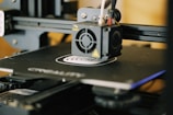 A close-up view of a 3D printer in action, focusing on the extruder head and the printed object forming on the print bed. The printer’s mechanical components and frame are visible, indicating its active status.