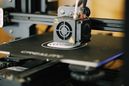 A close-up view of a 3D printer in action, focusing on the extruder head and the printed object forming on the print bed. The printer’s mechanical components and frame are visible, indicating its active status.