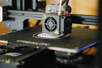 A close-up view of a 3D printer in action, focusing on the extruder head and the printed object forming on the print bed. The printer&rsquo;s mechanical components and frame are visible, indicating its active status.