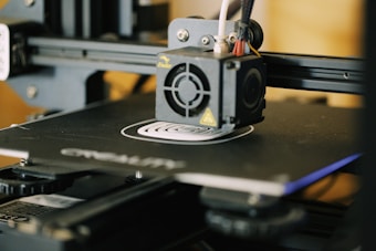 A close-up view of a 3D printer in action, focusing on the extruder head and the printed object forming on the print bed. The printer’s mechanical components and frame are visible, indicating its active status.