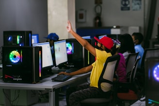 Several children are seated at a row of computers in a room, with one child raising their hand. They wear masks and colorful clothing, and the computers have LED lighting.