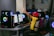 Several children are seated at a row of computers in a room, with one child raising their hand. They wear masks and colorful clothing, and the computers have LED lighting.