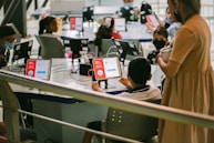 Several children are seated around a table, each using a tablet device with educational content displayed. An adult stands nearby observing the activity. The setting appears to be a classroom or learning environment, with the focus on digital learning.
