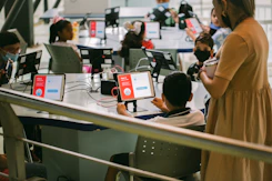 Several children are seated around a table, each using a tablet device with educational content displayed. An adult stands nearby observing the activity. The setting appears to be a classroom or learning environment, with the focus on digital learning.