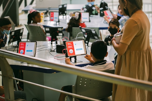 Several children are seated around a table, each using a tablet device with educational content displayed. An adult stands nearby observing the activity. The setting appears to be a classroom or learning environment, with the focus on digital learning.