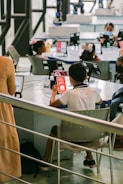 A group of children is seated at tables in a modern classroom or learning center, using tablets and wearing face masks. The environment is bright and airy with large windows and advanced equipment around the room. A teacher or instructor is present, providing guidance.