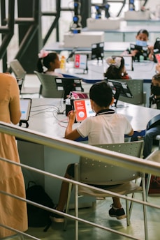 A group of children is seated at tables in a modern classroom or learning center, using tablets and wearing face masks. The environment is bright and airy with large windows and advanced equipment around the room. A teacher or instructor is present, providing guidance.
