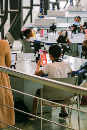 A group of children is seated at tables in a modern classroom or learning center, using tablets and wearing face masks. The environment is bright and airy with large windows and advanced equipment around the room. A teacher or instructor is present, providing guidance.